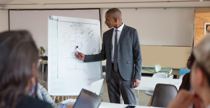 Confident speaker talking and pointing at whiteboard. Group of employees discussing ideas during presentation of new project at briefing. Business meeting concept