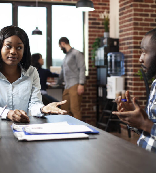 Young black woman explaining her work experiences and expectations to professional hiring manager. Male recruiter and female candidate engage in a formal discussion in a modern office.